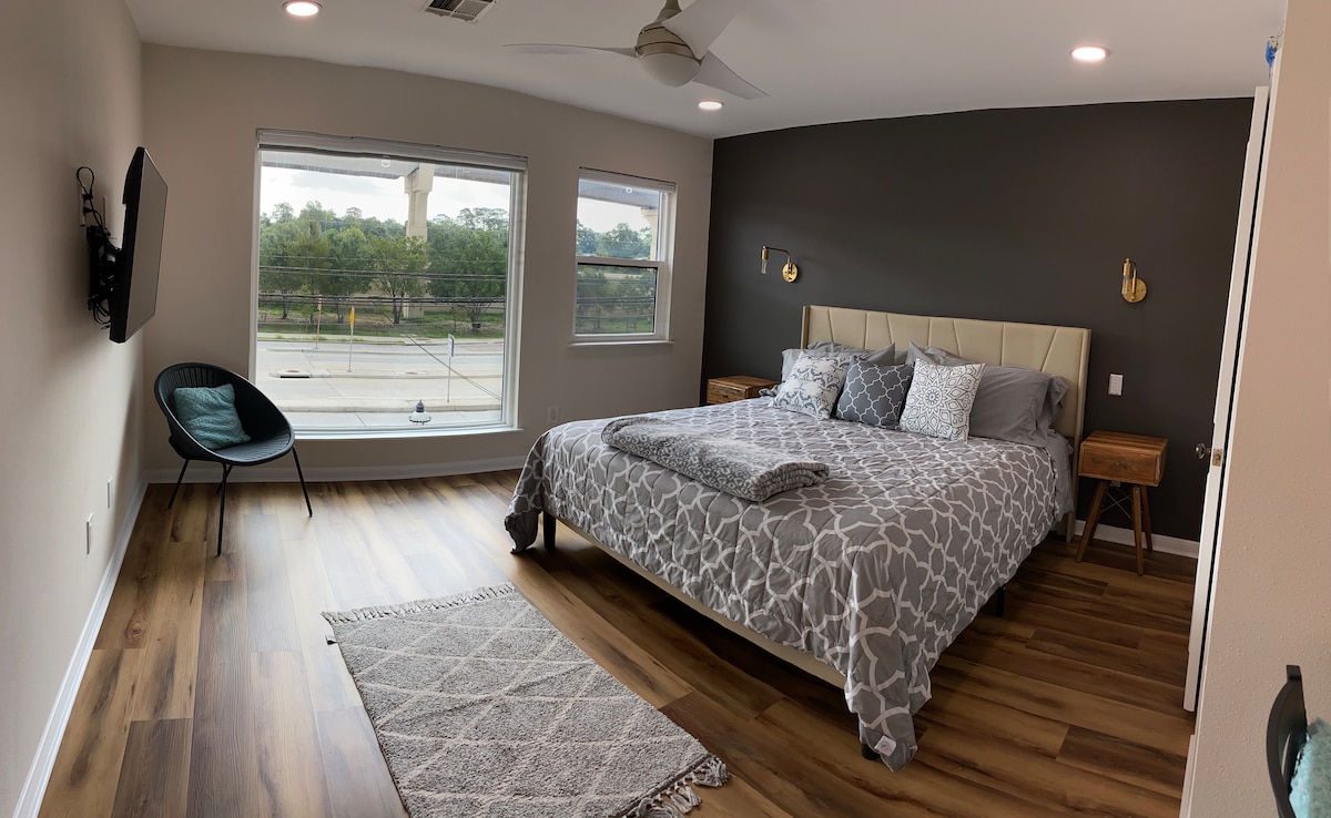 A spacious bedroom is showcased with a comfortable king size bed adorned with a neutral-patterned duvet. Two bedside tables flank the bed, and a light grey area rug lies on the warm hardwood floor. Large windows allow natural light to illuminate the room, featuring a view of greenery.