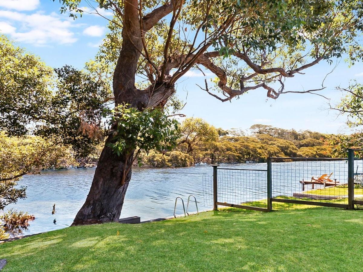 A lush green lawn leads to the tidal creek, framed by trees and shrubbery. In the foreground, a set of steps descends towards the water, while a wooden lounge chair is positioned nearby, offering a serene view of the natural surroundings.