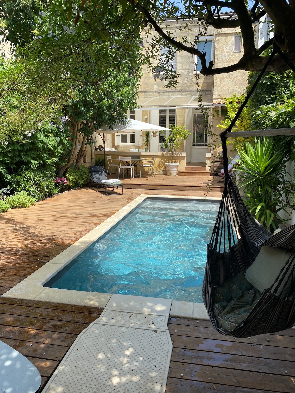 A serene garden space features a small pool surrounded by wooden decking. Lush greenery frames the area, with a dining set visible nearby under a shaded umbrella. A hammock is suspended between two trees, inviting relaxation in this tranquil outdoor setting.