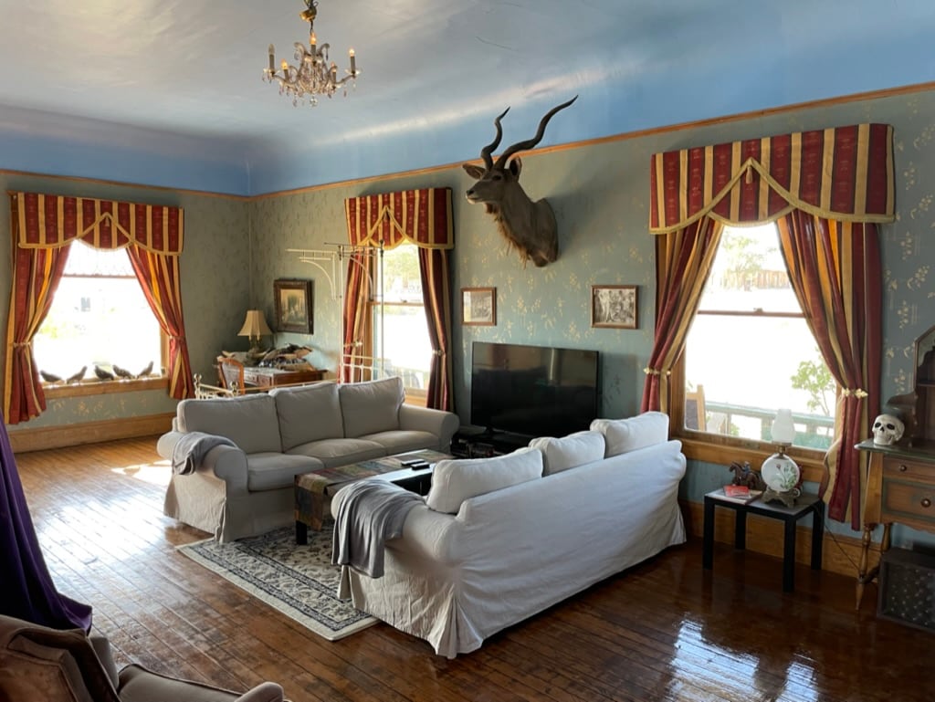 A spacious living room features comfortable seating with a sectional sofa and an armchair, complemented by a large TV. Natural light enters through two windows, draped in patterned curtains. A decorative chandelier hangs from the ceiling, while a mounted deer head adds a unique character to the space.