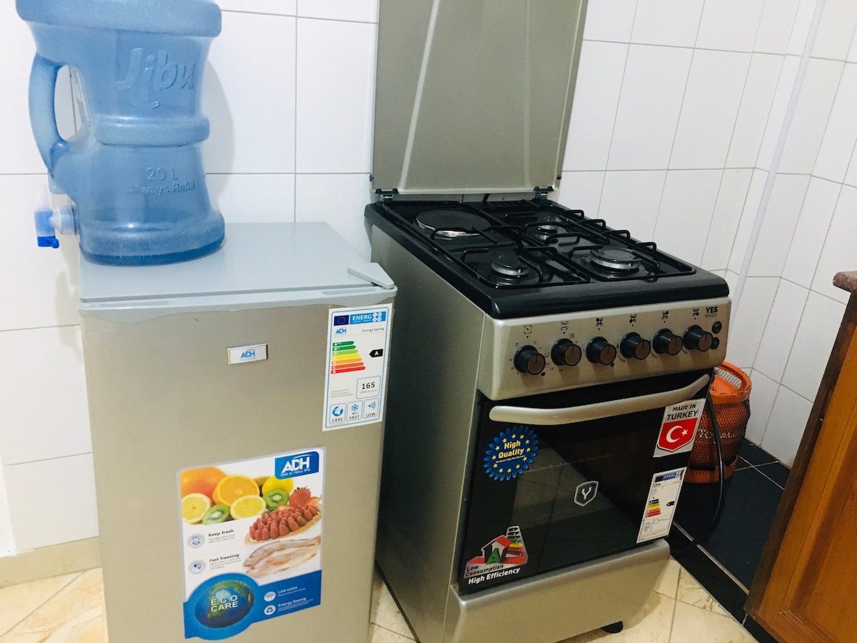 The kitchen features a compact refrigerator and a stainless steel gas stove with four burners. A water dispenser is placed on top of a counter. White tiled walls provide a clean backdrop, while a decorative mat adds a touch of color to the floor.