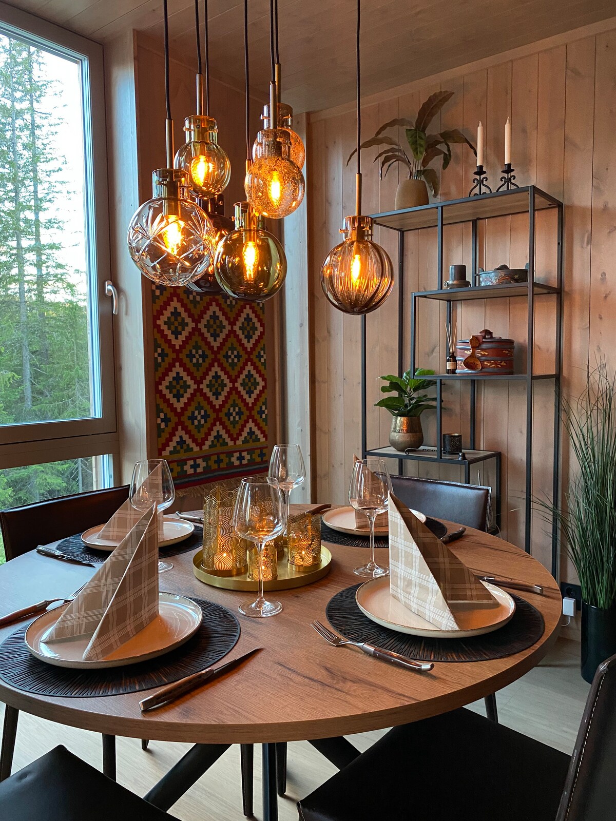 A round wooden dining table is set for a meal, featuring stylish plates and glasses, with triangular fabric napkins elegantly arranged. Unique pendant lights hang above, providing warm illumination. A decorative shelving unit showcases plants and decorative items, enhancing the cozy atmosphere of the dining area.
