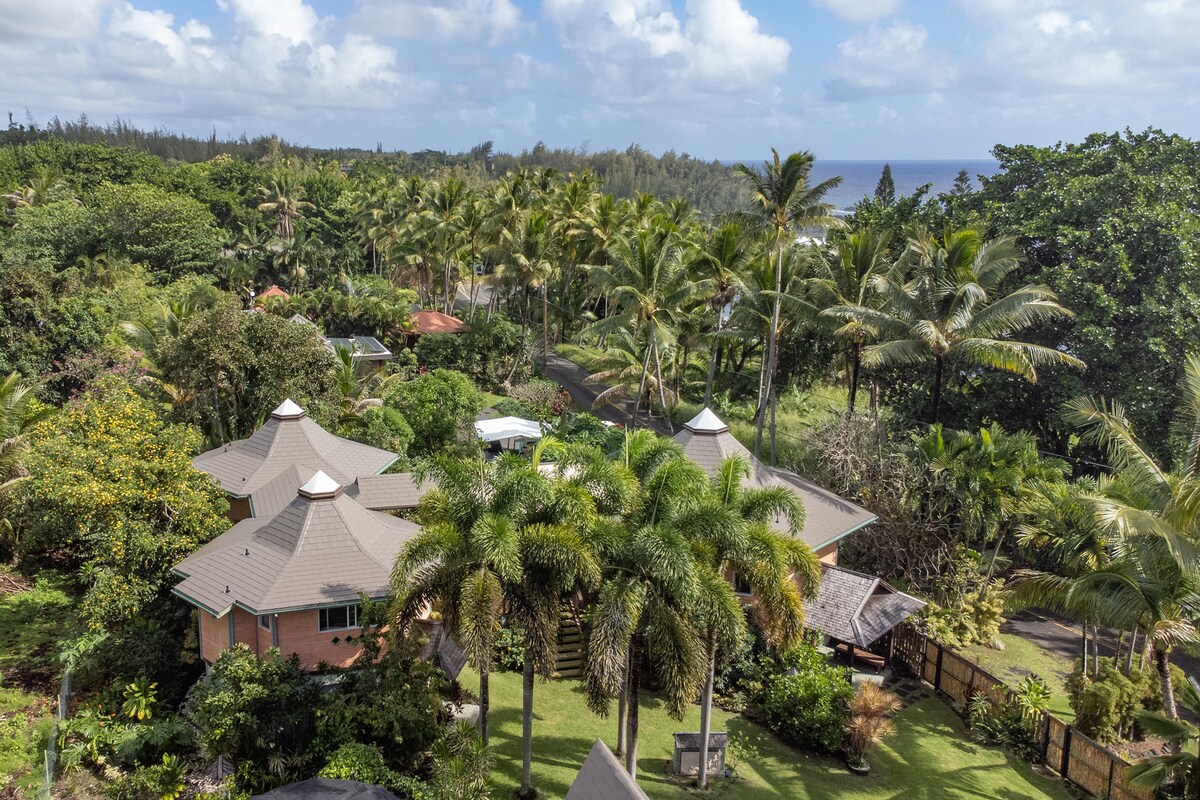An aerial view captures the Balinese-style residence nestled among lush greenery and palm trees. The ocean can be seen in the distance, complementing the vibrant landscaping that surrounds the home. The unique architecture features multiple roofs and an inviting outdoor space.
