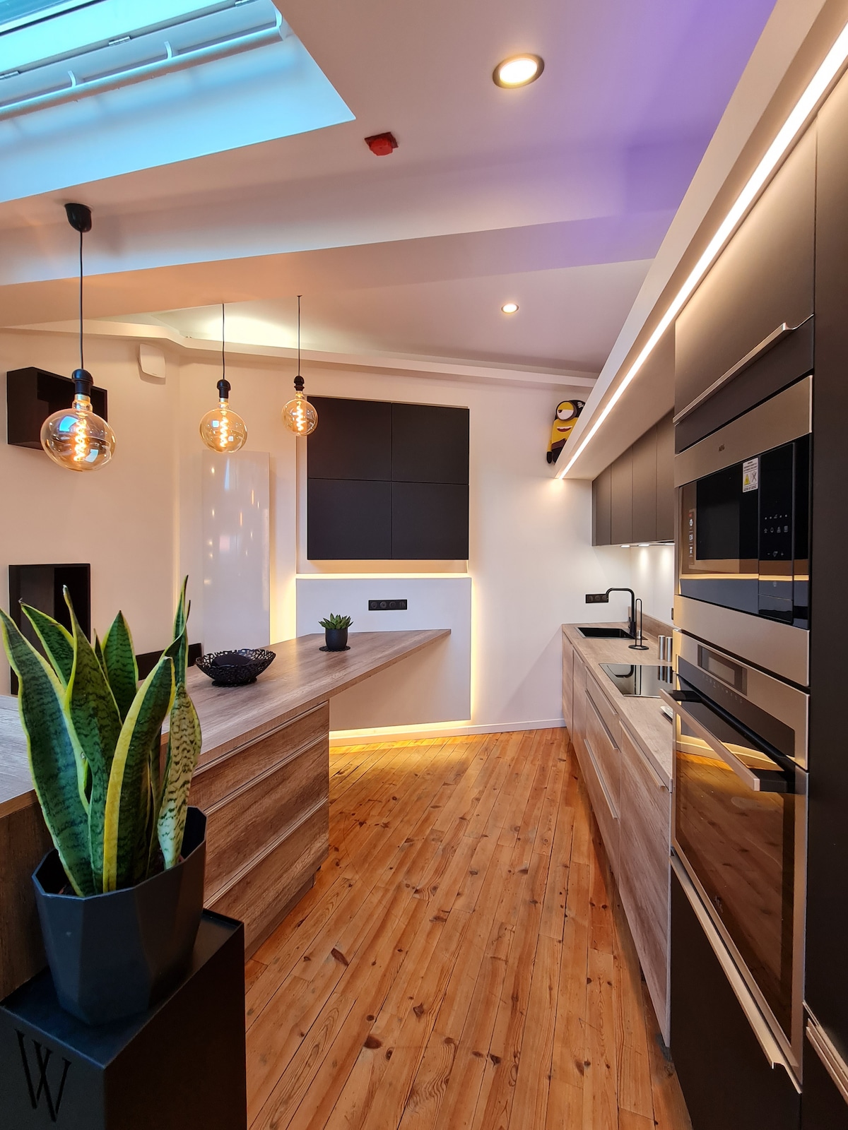 A modern kitchen features sleek black cabinetry and stainless steel appliances, complemented by warm wood accents. Soft lighting from pendant fixtures illuminates the space. A potted plant adds a touch of greenery, enhancing the kitchen's contemporary design.