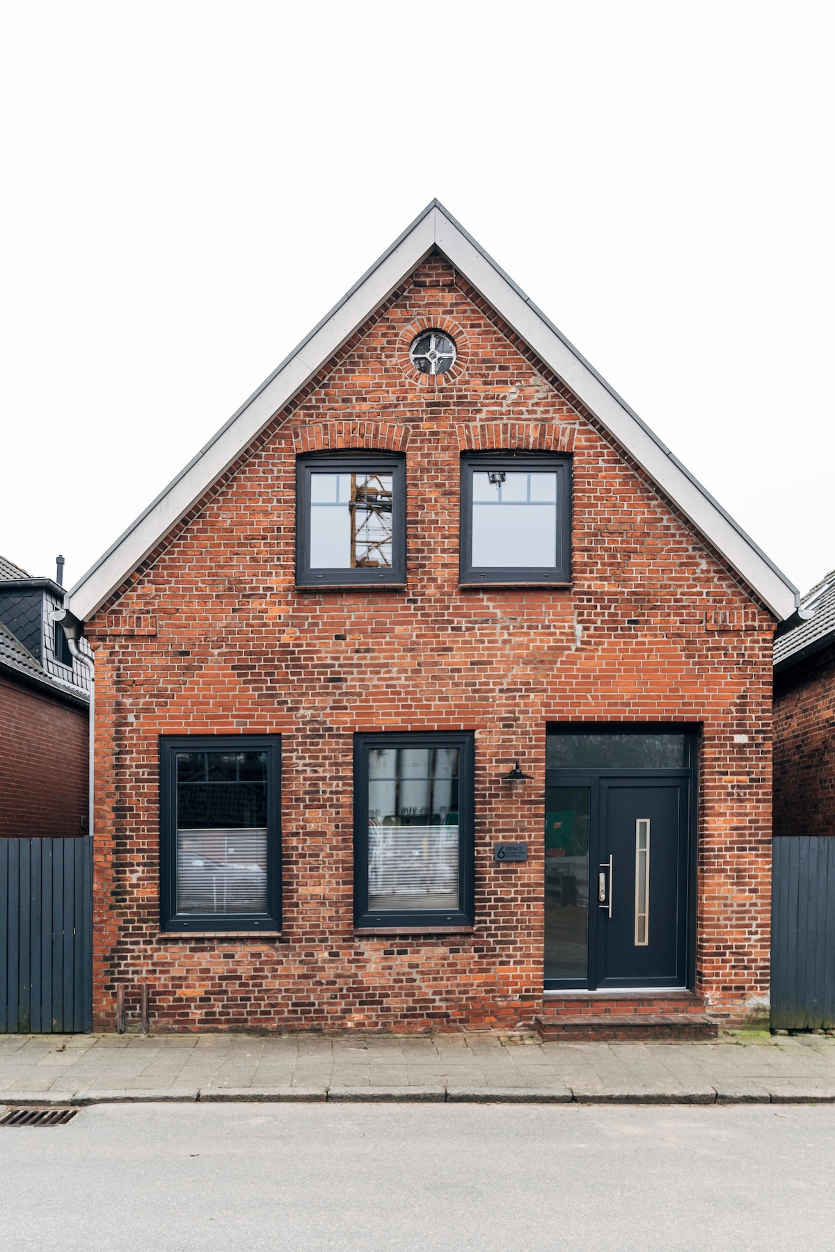 The exterior of a charming two-story brick house is presented, featuring a distinct triangular gabled roof. Three windows are symmetrically arranged, allowing natural light to enter. A modern front door is positioned centrally, enhancing the building's welcoming appearance. A cobblestone pathway leads to the entrance.