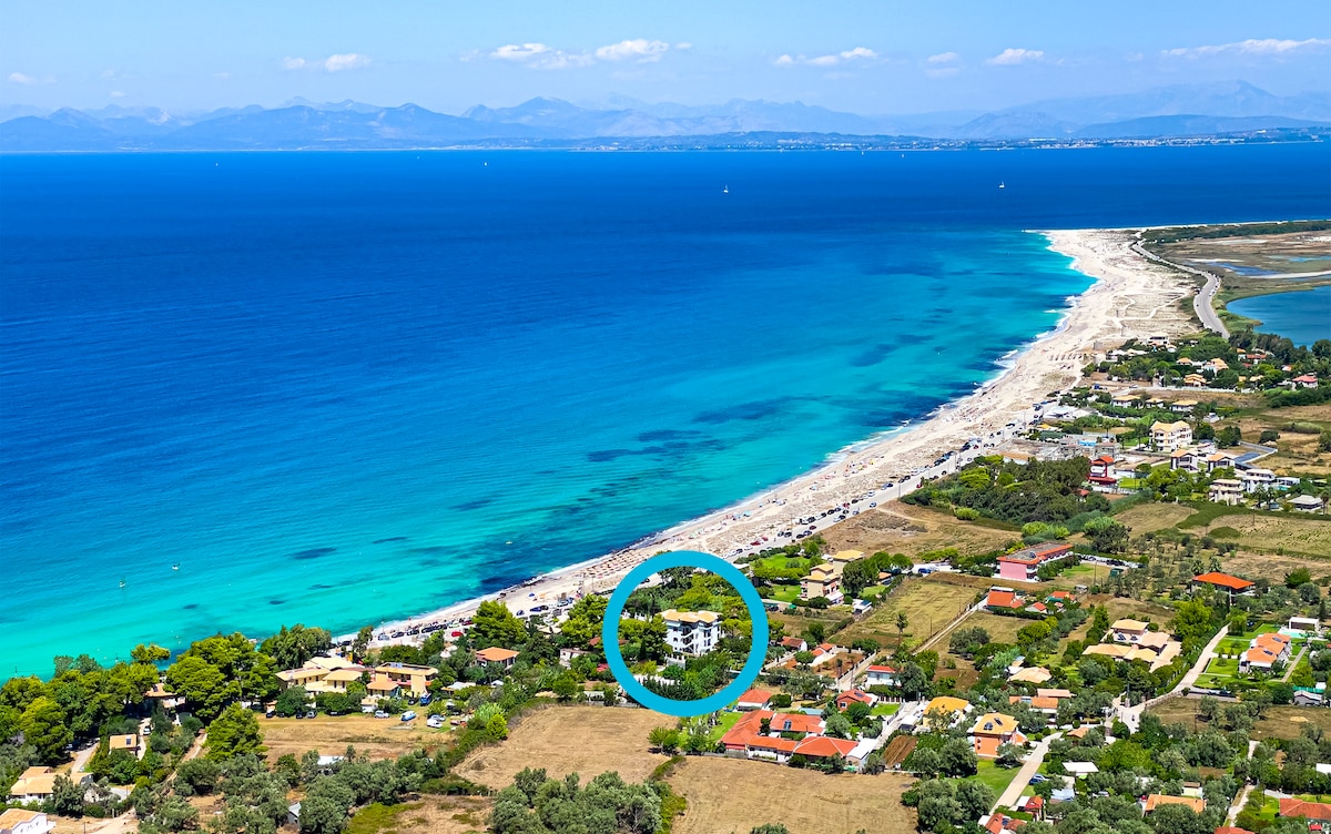 An aerial view captures the location of the house near a sandy beach, with clear blue waters of the Ionian Sea stretching out to the horizon. Surrounding greenery and residential areas are visible, providing a sense of the tranquil environment and the proximity to the beach.