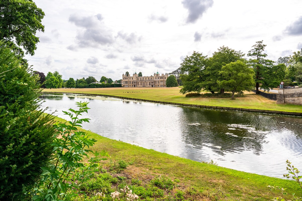 The image captures a serene view of a large estate situated across a calm body of water. Lush greenery lines the banks, complemented by manicured lawns leading to the historic building. Fluffy clouds drift in a light gray sky, accentuating the tranquil setting.