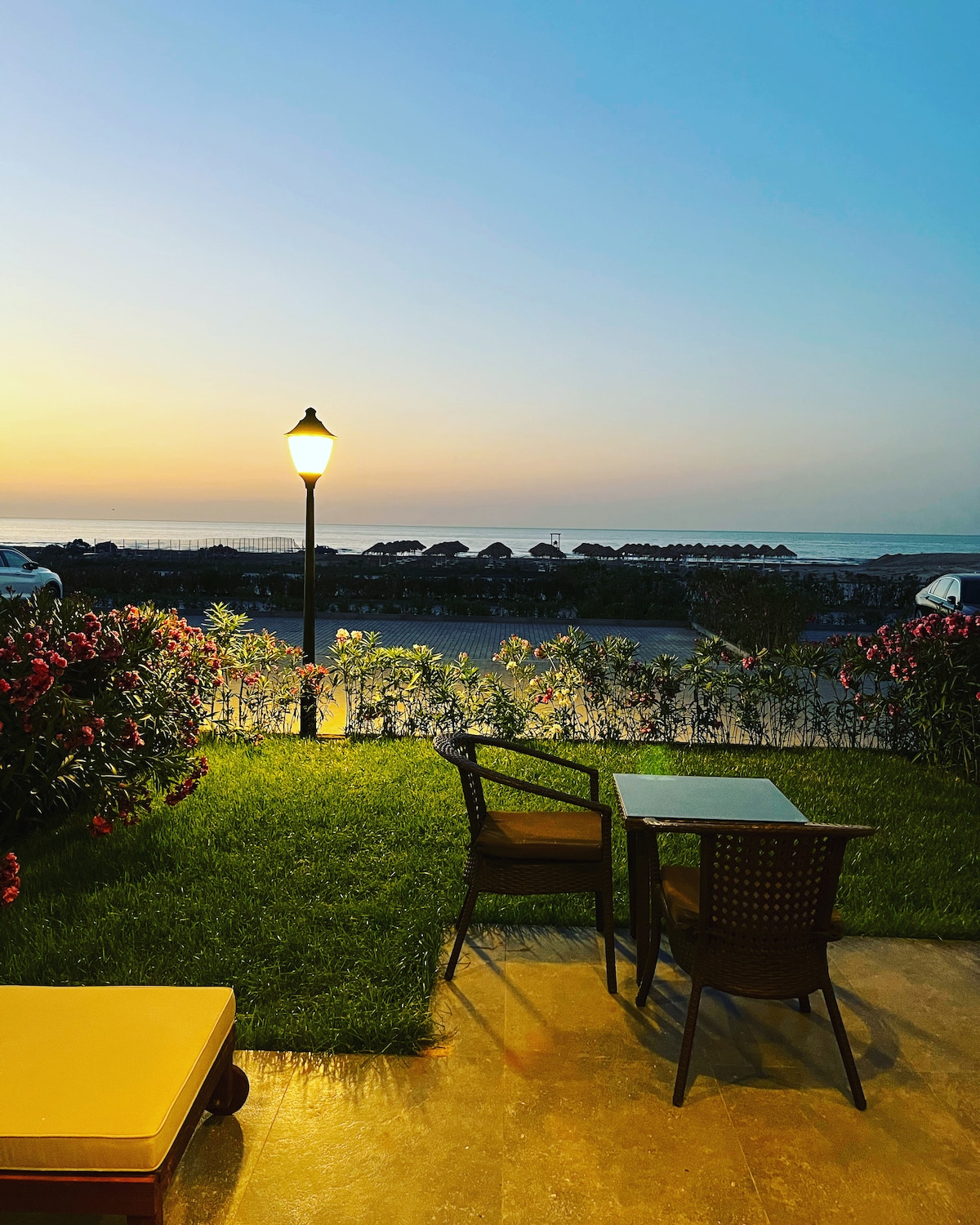 A tranquil outdoor seating area features a small table and two chairs positioned on lush green grass. A decorative lamp stands nearby, casting a warm glow as the sun sets over the calm water, creating a peaceful ambiance.