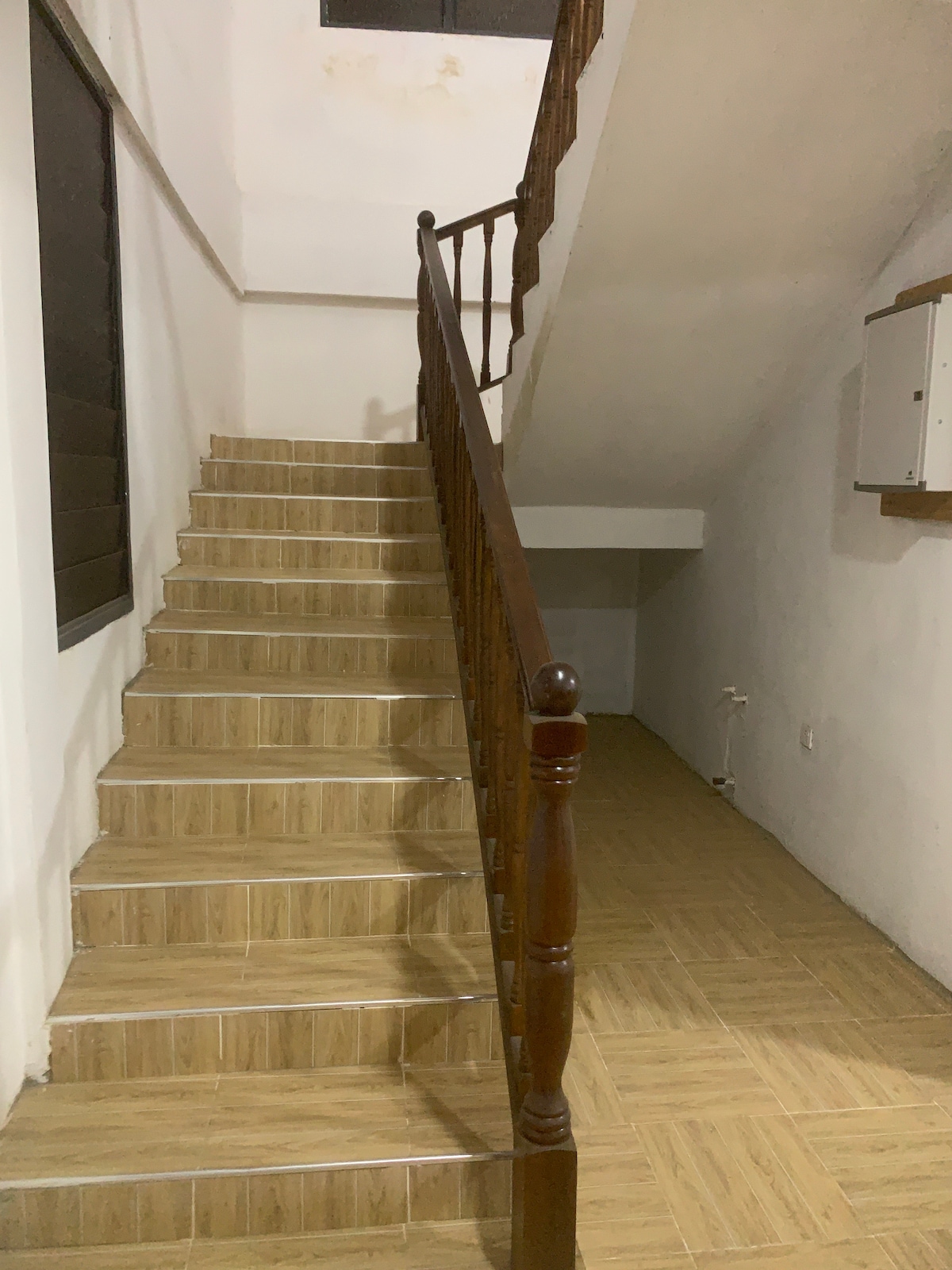 A staircase with wooden railings leads upwards, featuring beige tiled steps that complement the light-colored walls. Natural light filters through a nearby window, illuminating the area. The landing at the top is open and spacious, with a minimalistic design.