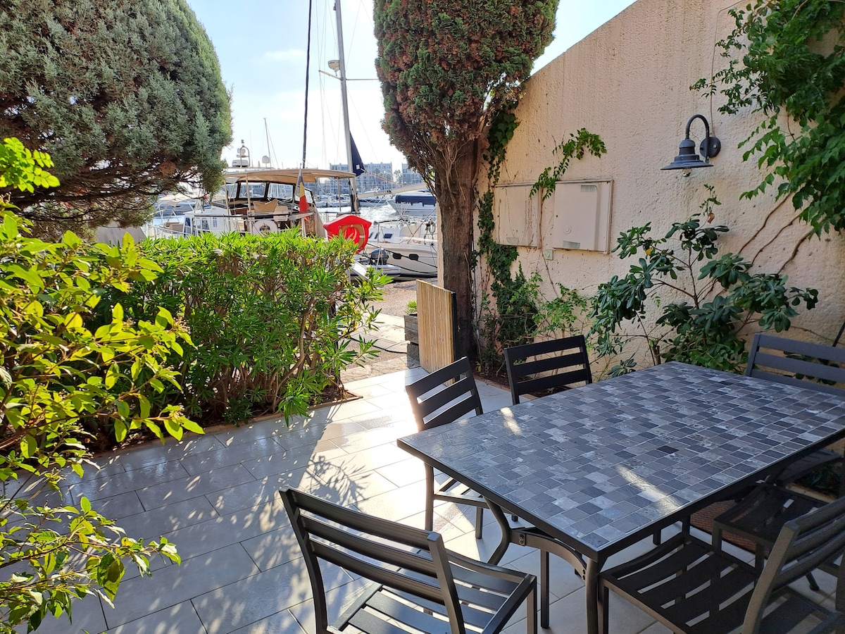 A patio area features a large dining table surrounded by several black chairs, set against a backdrop of lush greenery. The terrace provides a view of the nearby marina, with boats moored in the water visible through the foliage.