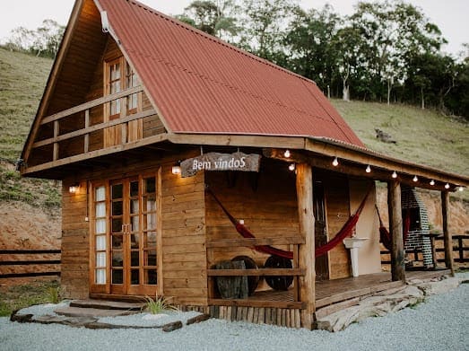 The cabin features a rustic wooden exterior with a red roof, surrounded by greenery. A welcoming sign is displayed above the door. Hammocks are hung on the porch, providing a comfortable space to relax and enjoy the serene landscape.