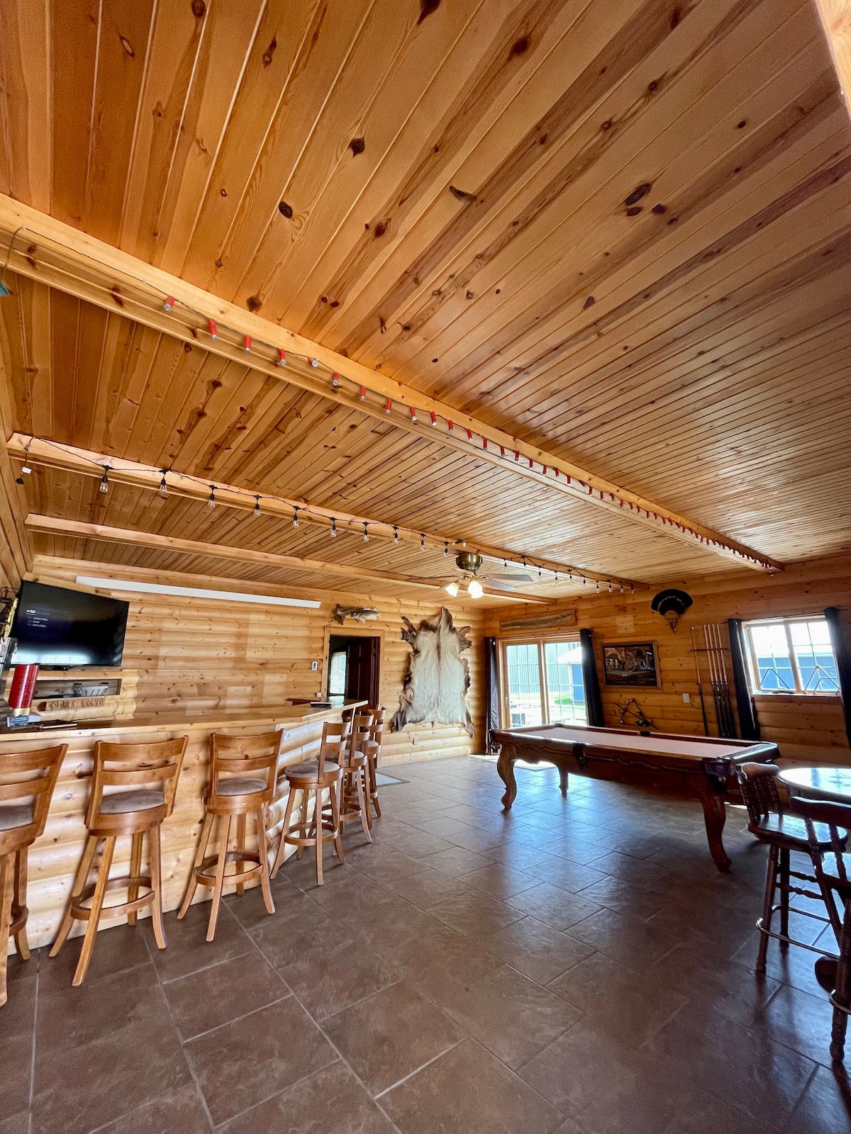 The spacious common area showcases wooden paneling with a high ceiling, featuring a bar with several stools and a pool table. A large television is mounted on one wall, while decorative elements enhance the rustic charm. Natural light fills the space through windows along the far wall.