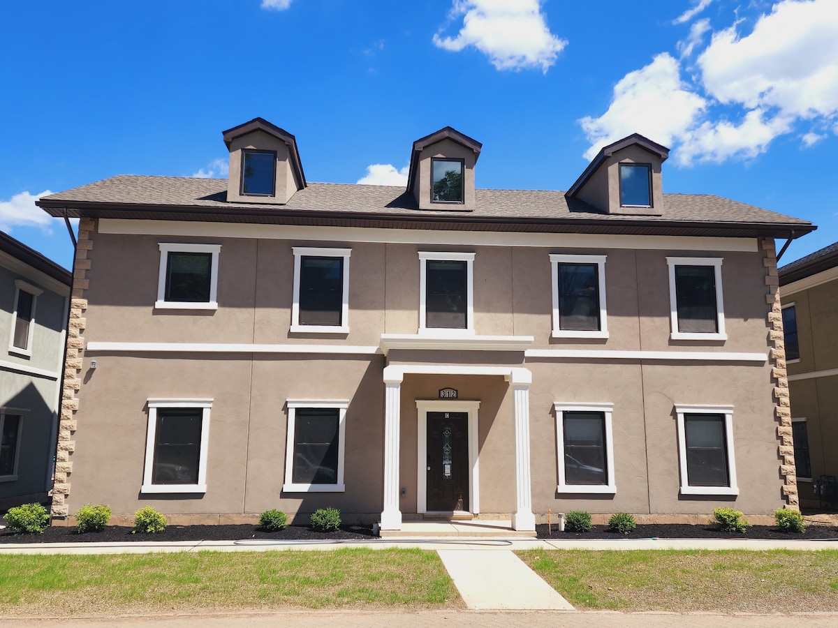 The exterior of the newly built townhome is presented with a modern design, featuring a symmetrical façade with multiple windows. A central entryway is framed by a small portico, and neatly landscaped plants are positioned along the front. The building is set against a bright blue sky.