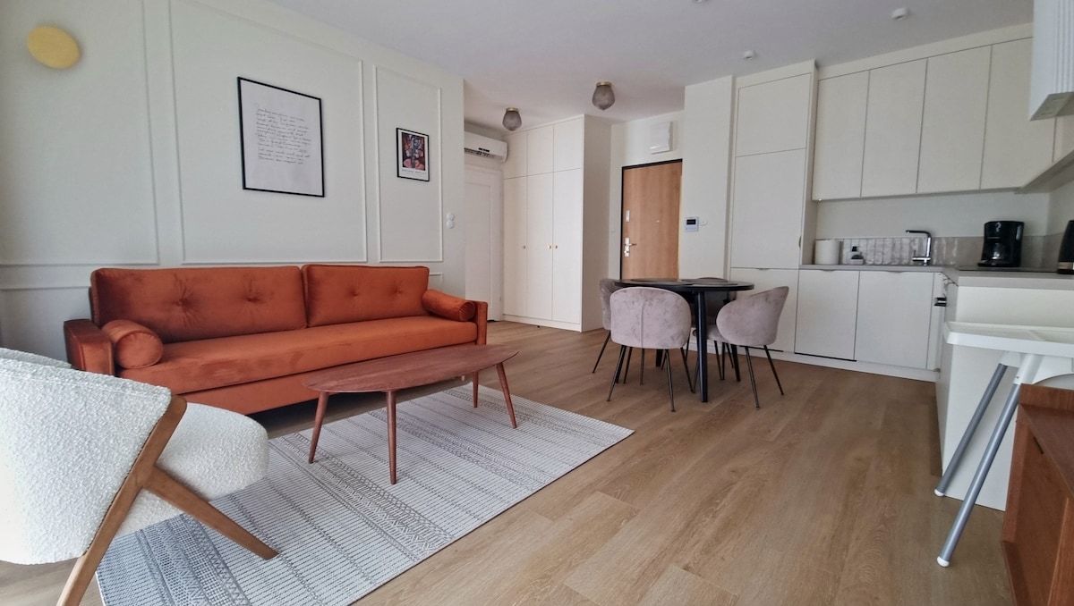 A modern living space features a vibrant orange sofa and a stylish wooden coffee table, complemented by a round dining table and four plush chairs. The kitchen area is equipped with essential appliances, and natural light fills the room through large windows. A simple rug adds texture to the wood flooring.