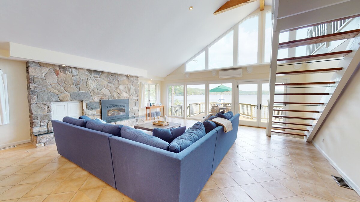 A spacious living area features a large sectional sofa facing a stone gas fireplace, with exposed wood beams overhead. Floor-to-ceiling windows provide unobstructed views of the lake. A staircase leads to the upper level, while outdoor seating is visible on the deck.