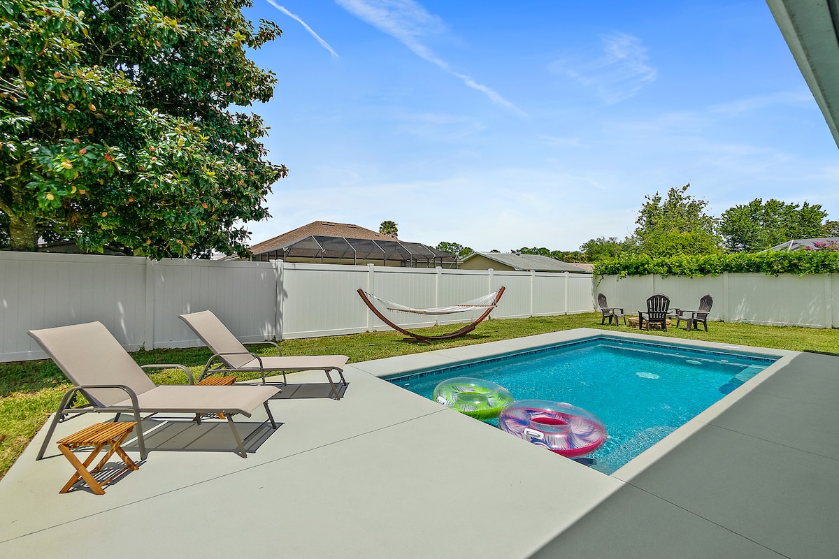 A well-maintained outdoor space features a heated saltwater pool surrounded by a large patio. Lounge chairs are positioned for relaxation, while colorful inflatables float in the water. A comfortable hammock and seating area are visible, providing options for leisure and enjoyment.