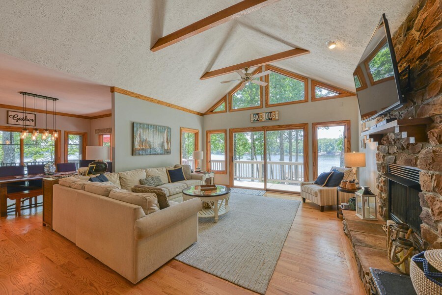 The open concept living area features a comfortable seating arrangement with a large sectional sofa positioned around a coffee table. Large windows offer views of the lake, while a stone fireplace adds a cozy touch. Natural light fills the space, highlighting wooden beams on the ceiling.