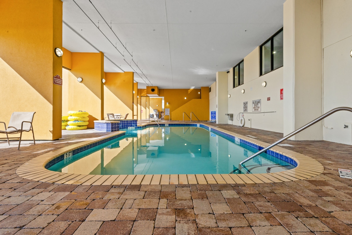 An indoor pool area features a clear, inviting pool surrounded by warm-toned walls. Lounge chairs line one side, while bright yellow floatation devices are stacked nearby. Natural light floods the space, highlighting the tiled flooring and creating a relaxing environment.