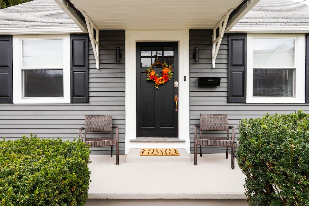 The entrance to the home is shown, featuring a solid black door adorned with a seasonal wreath. Two wicker chairs sit on either side of the door under awnings, with neatly trimmed shrubs lining the pathway for a welcoming look.