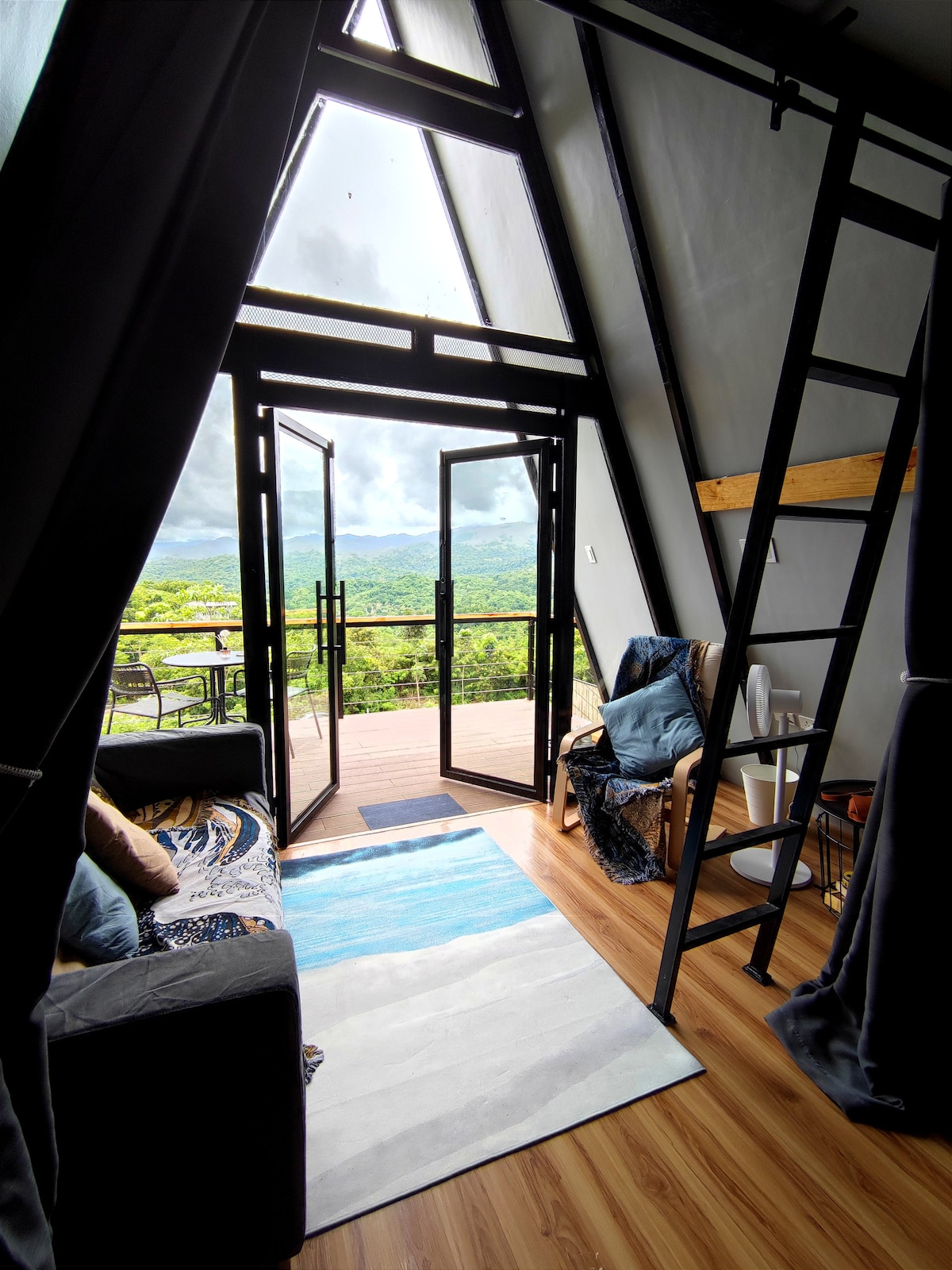 An inviting entryway features an A-frame design with large glass doors that open to reveal breathtaking mountain views. A cozy seating area is set against the vibrant backdrop, complemented by a soft rug and decorative throw pillows. A wooden ladder leads to the loft space.