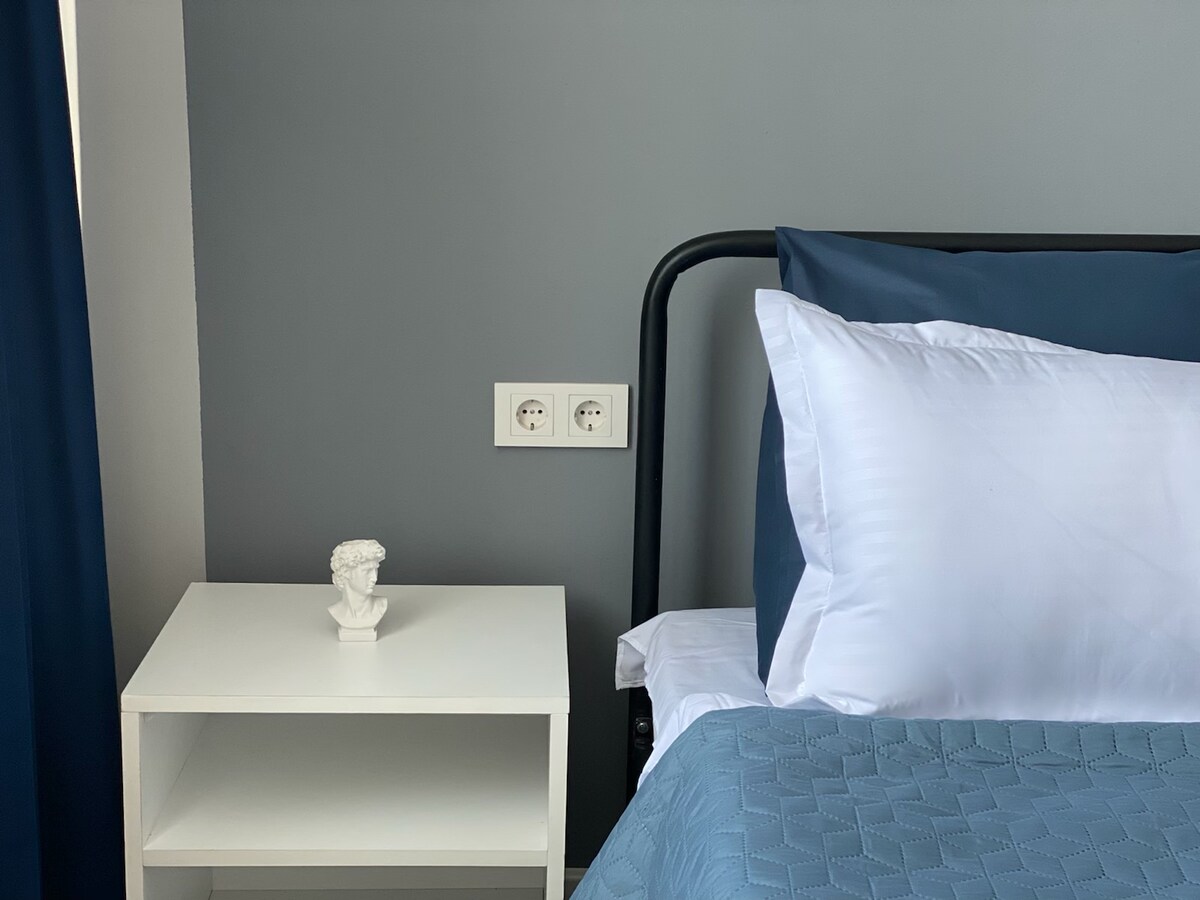 A minimalist bedroom setting features a sleek black metal bed with crisp white pillows and a textured blue blanket. A small white bedside table is positioned next to the bed, displaying a decorative figurine. The walls are painted in a calm gray tone.