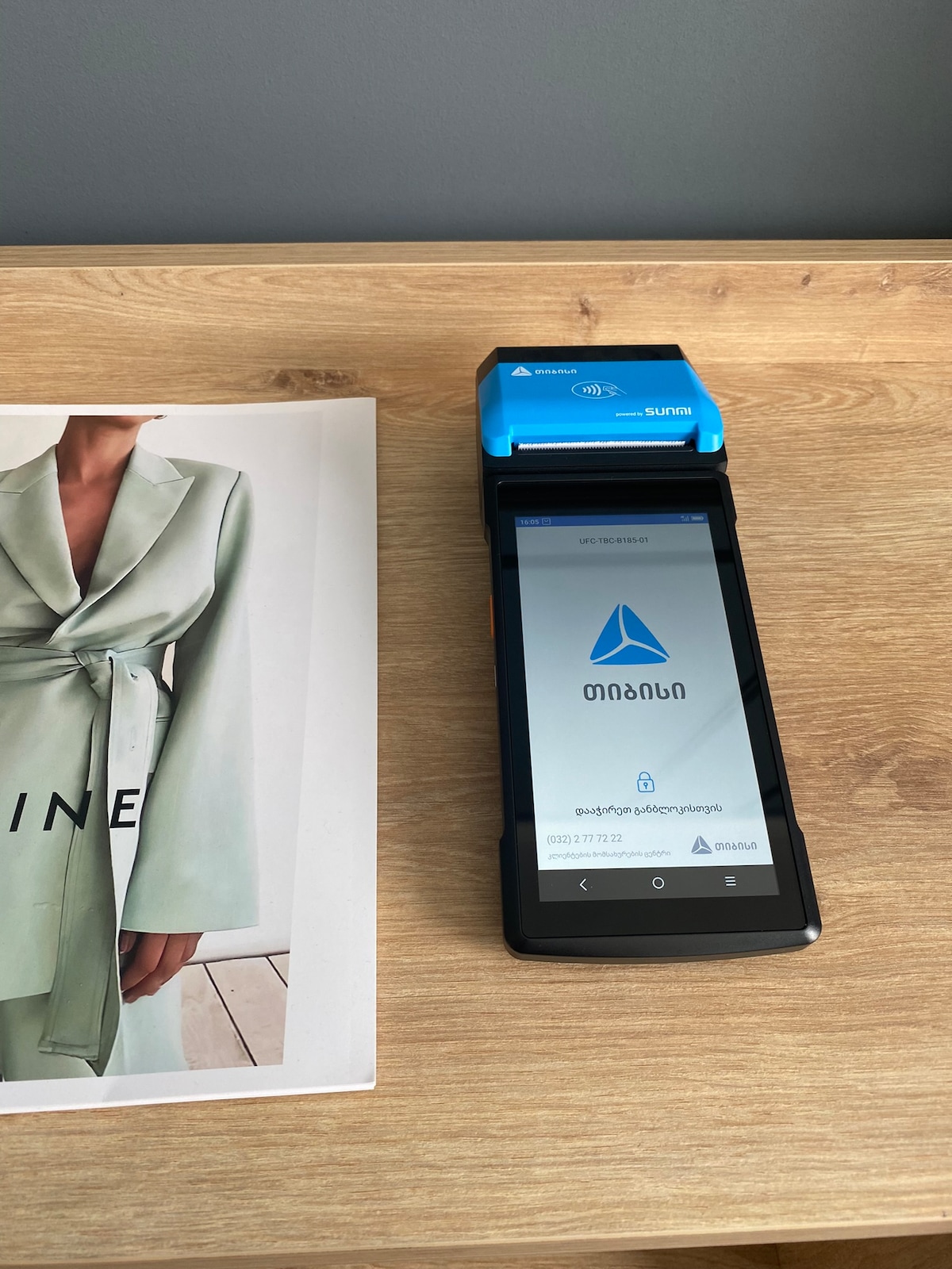 A wooden tabletop displays a sleek mobile device and a compact payment printer. The device screen reads a payment interface, while a fashion magazine with a soft green cover lays beside it, providing a contemporary and functional workspace.