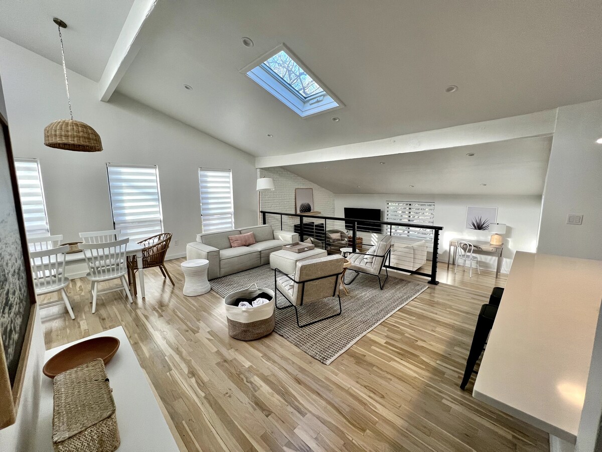An airy living space is showcased with a mix of comfortable seating options, including a sectional sofa and chairs. Natural light enters through a skylight and large windows adorned with sheer coverings. A dining area sits adjacent, featuring a table with seating. Light wood flooring complements the decor.