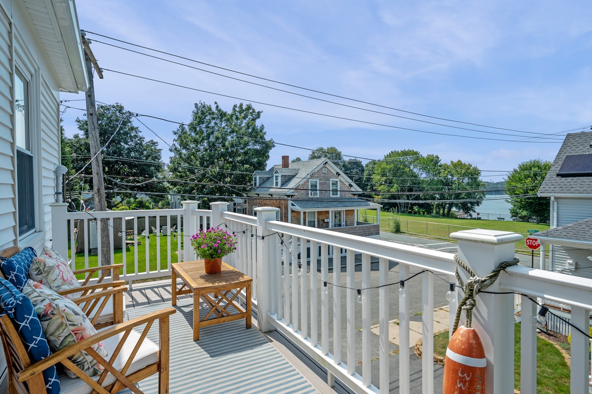 An outdoor deck features two wooden chairs with colorful cushions and a small table, complemented by a blooming flower arrangement. Scenic views of the Danvers River and surrounding greenery are visible, with light blue skies overhead and nearby residential structures in the background.