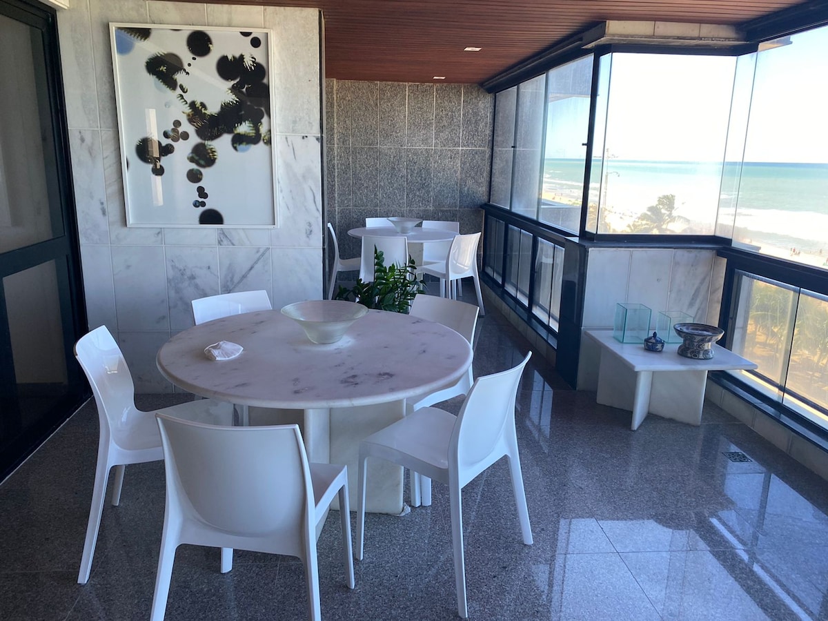A spacious balcony features a round table surrounded by white chairs, offering a clear view of the sea. Large windows illuminate the area, while a decorative plant adds a touch of greenery. The marble flooring enhances the elegant atmosphere.