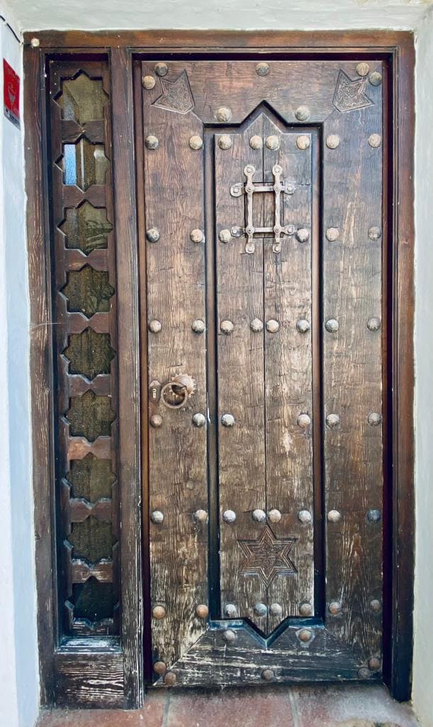 An intricately designed wooden door features a rich, dark finish adorned with decorative metal studs and a unique emblem. To the side, a narrow window with geometric patterns allows natural light to filter through, enhancing the architectural charm of the entrance.