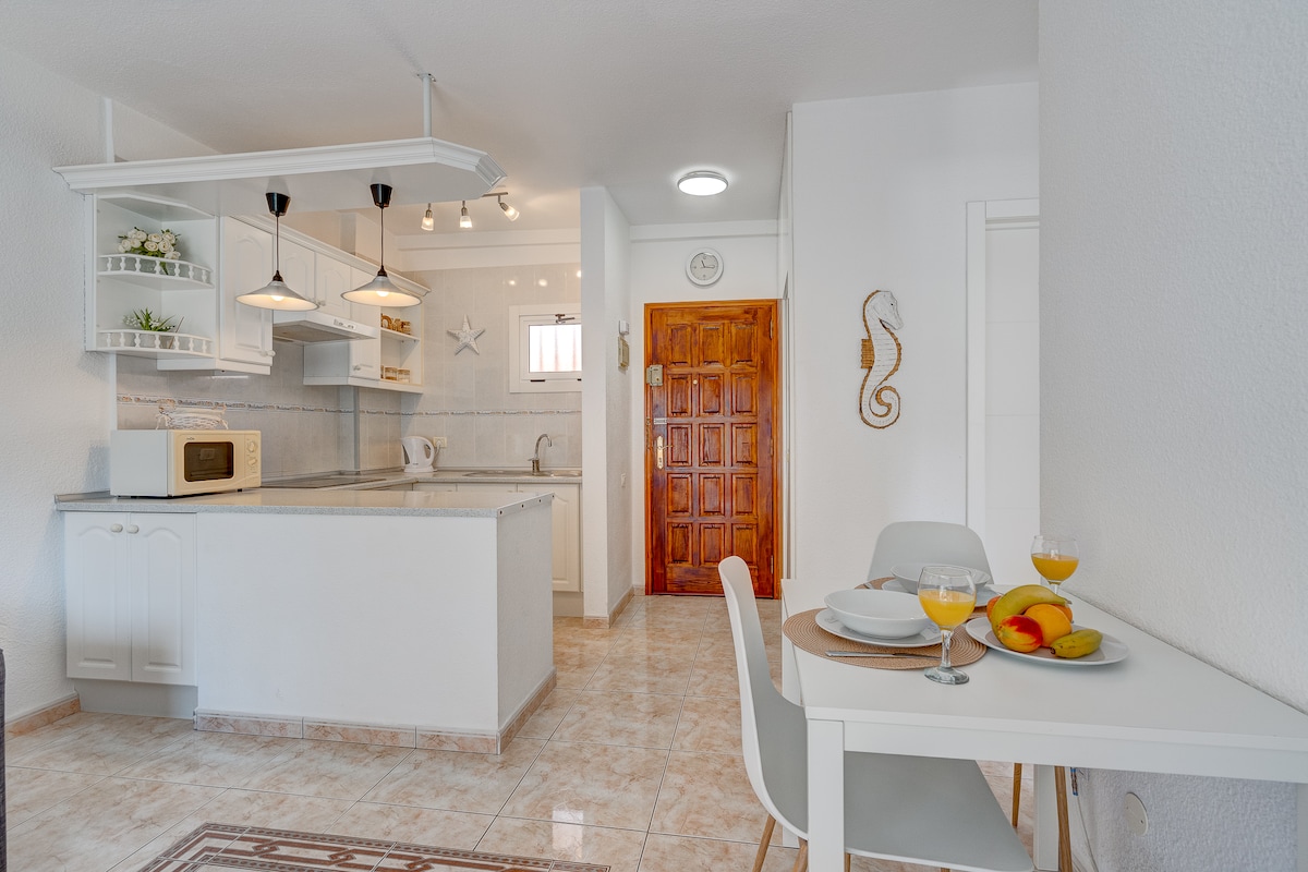 A bright kitchen area is equipped with a microwave and features a dining table set for two. Large windows allow natural light to fill the space, while light-colored cabinets and countertops maintain a fresh and inviting ambiance.