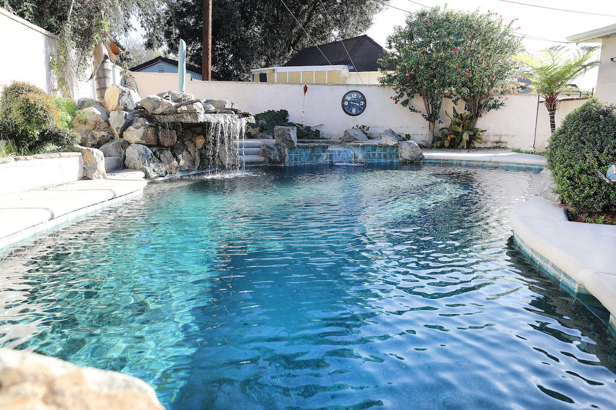 A serene salt-water pool is surrounded by natural stone features and lush greenery, with a cascading waterfall gently flowing into the water. The pool is designed with smooth curves and a blend of light blue and clear water, providing a refreshing retreat.