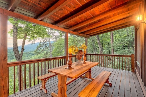A wooden deck features a rustic table and benches, surrounded by tall trees and expansive mountain views. Soft lighting is provided by a wall-mounted fixture, enhancing the serene environment. The natural wood tones and large beams contribute to a cozy, secluded setting.