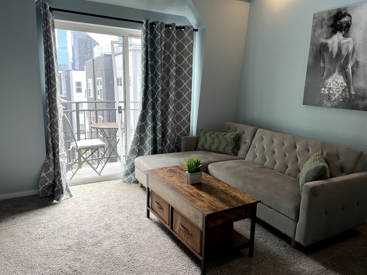 A comfortable living area features a grey sectional sofa and a wooden coffee table. Natural light filters in through a sliding door, which leads to a small balcony. Light curtains and plush carpet enhance the cozy ambiance of the space.