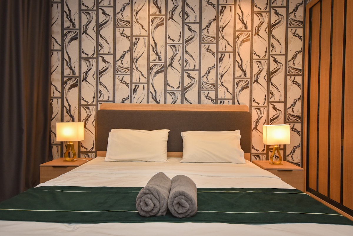 A well-appointed bedroom features a spacious bed with neutral bedding. Two bedside lamps are positioned on nightstands, providing soft lighting. Gray towels are neatly placed on the bed, complementing the patterned wallpaper that adds character to the space.