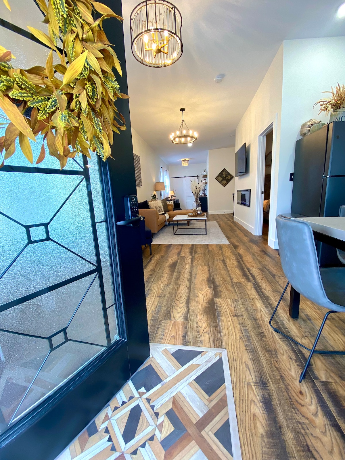 A view through the entryway showcases a hardwood floor leading into a well-lit living area. The hallway is adorned with decorative light fixtures, and the cozy seating area features a modern TV setup. A dining area is visible in the foreground, enhancing the inviting atmosphere.