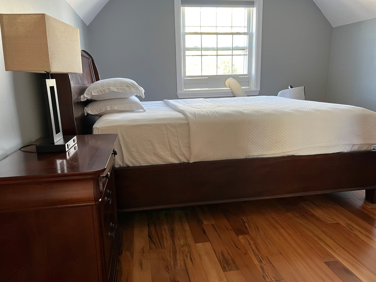 A well-arranged bedroom features a spacious bed with crisp white linens, positioned against a light gray wall. A wooden nightstand with a lamp is located beside the bed, and hardwood flooring adds warmth to the space. Natural light flows in through a window.