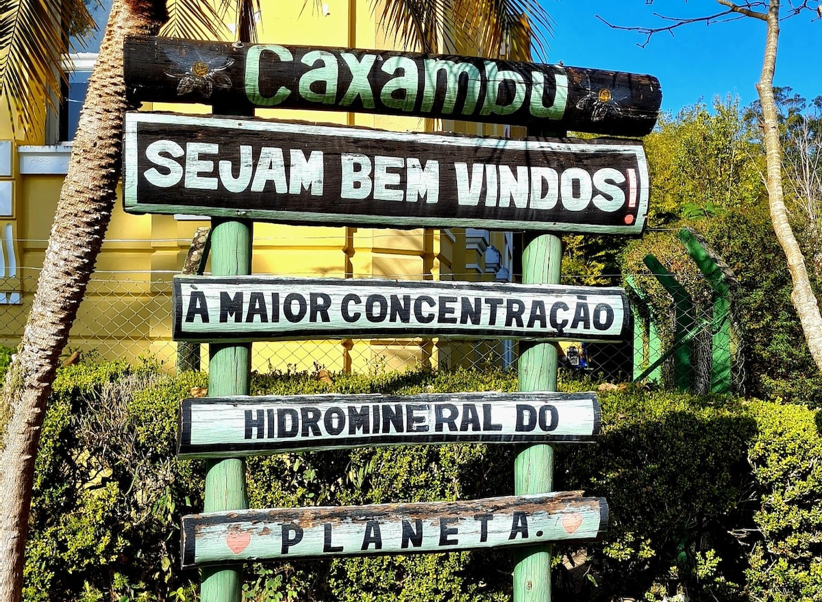 A welcoming sign at the entrance of Caxambu greets visitors with the phrase 'Sejam Bem Vindos!' The sign emphasizes Caxambu's distinction as home to the largest concentration of mineral water in the world, surrounded by greenery and palm trees.