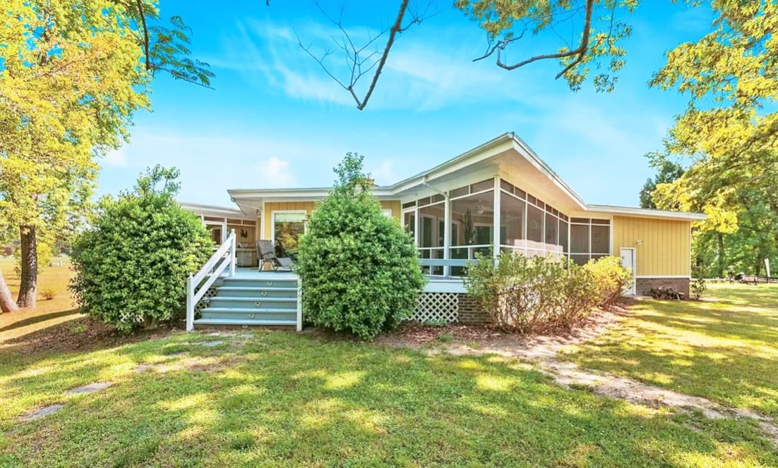 A spacious cottage is surrounded by landscaped green lawns and mature trees. The exterior features a screened-in porch, accessible via a set of stairs. Large windows reflect sunlight, suggesting a bright and airy atmosphere throughout the home.