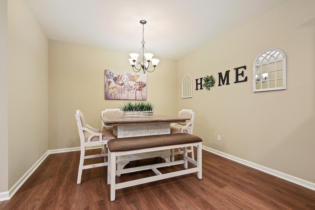 A dining area is presented with a long wooden table, surrounded by light-colored chairs and a bench. A chandelier hangs above, providing warm light. Decor includes a floral wall art piece and the word 'HOME' displayed on the wall, complemented by two decorative mirrors.