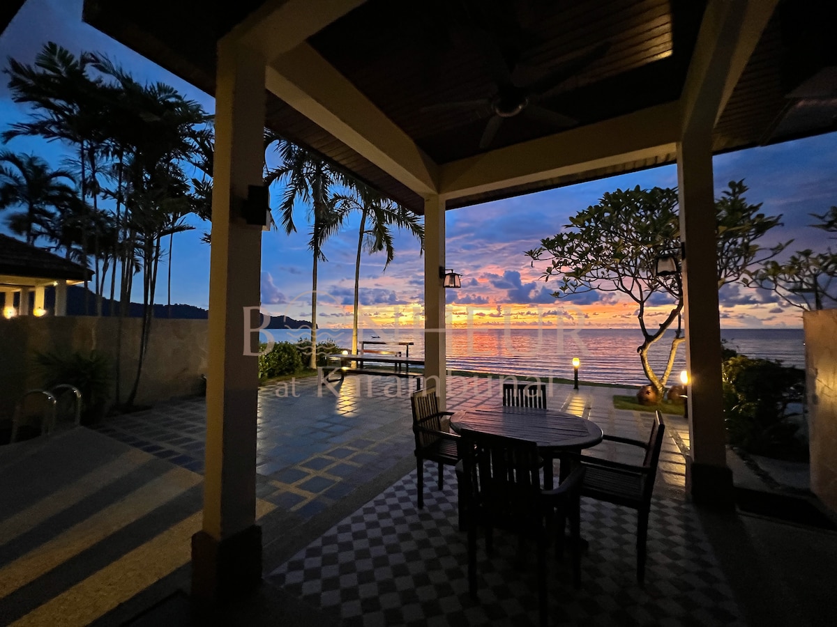 A covered patio area is featured, showing a dining table surrounded by chairs. The sunset casts soft colors across the ocean view, complemented by palm trees in the foreground. The space is illuminated, enhancing the tranquil ambiance of the beachfront setting.