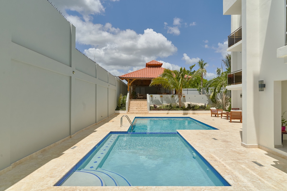 A sunlit outdoor area features two sparkling pools surrounded by a paved deck. A gazebo with a terracotta roof offers shaded seating, while tropical plants enhance the serene setting. The building's modern architecture provides a clean and inviting backdrop.