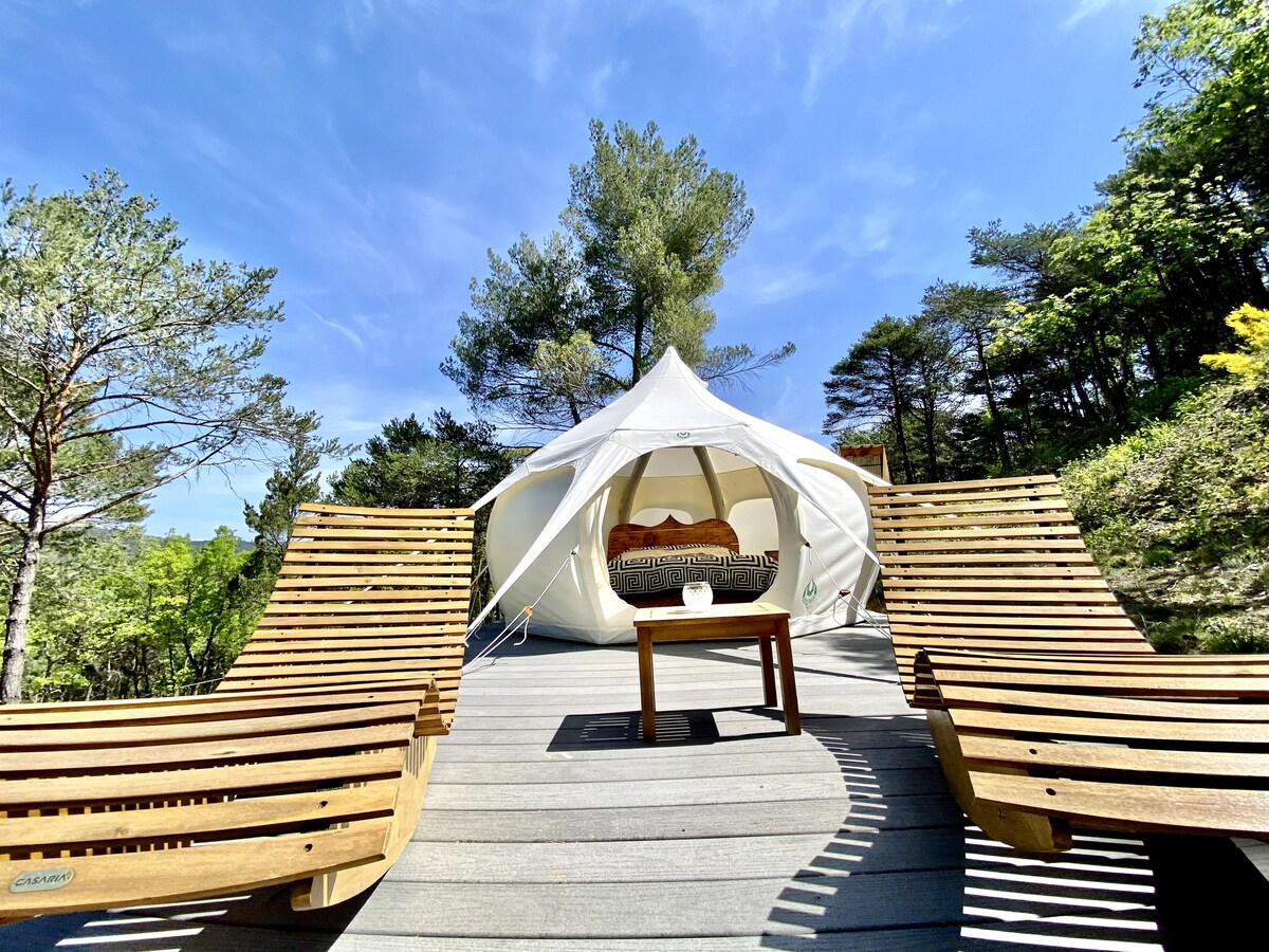 A white Lotus tent is positioned on a wooden deck, surrounded by lush green trees. Two wooden lounge chairs are angled toward a small table, inviting relaxation in a serene outdoor setting. The clear blue sky and sunlight enhance the tranquil atmosphere.