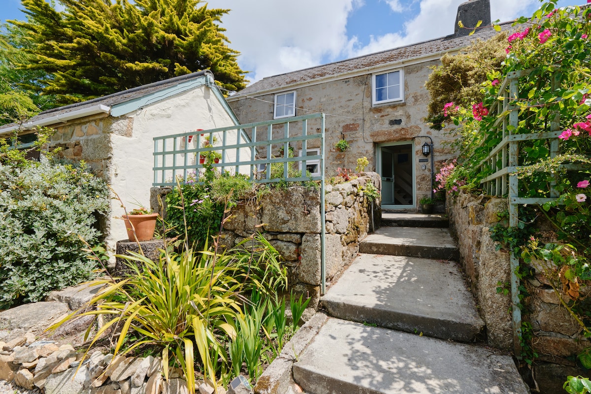 A stone cottage entrance is framed by lush greenery and colorful flowers. A stone pathway leads to the door, bordered by low stone walls and trellises. Sunlight filters through the surrounding plants, creating a welcoming atmosphere for guests.