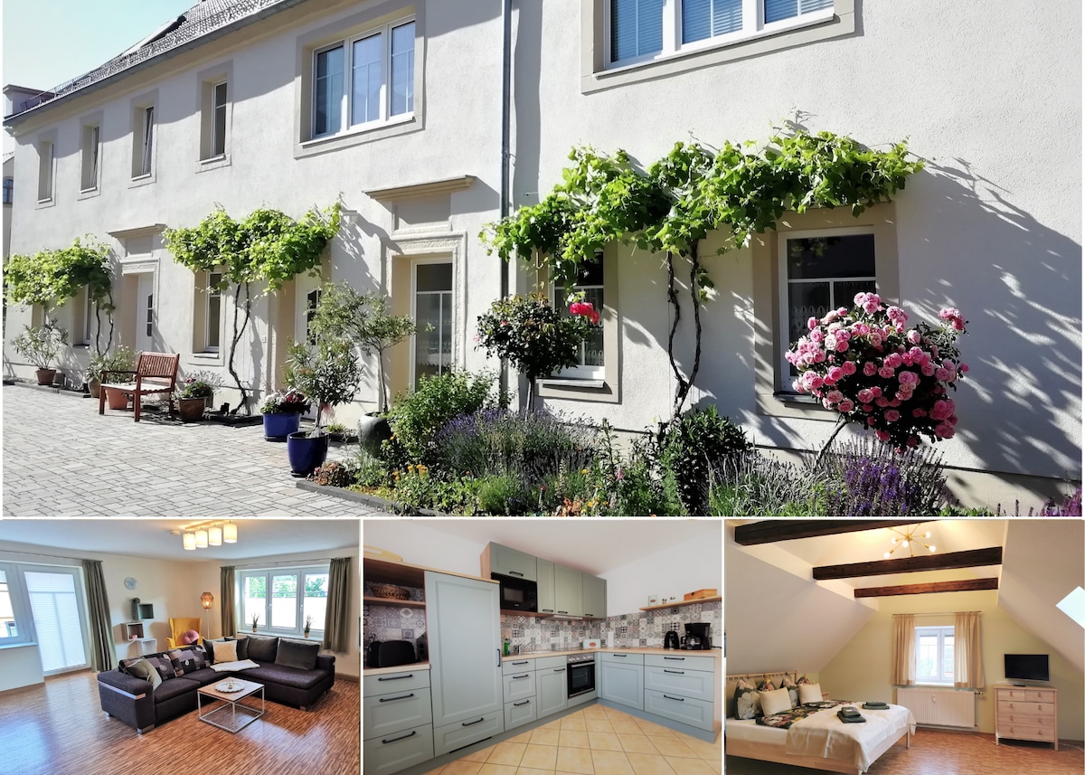 The exterior features a charming facade with window boxes adorned with blooming flowers and climbing vines. The interior combines a spacious living area with comfortable seating, a modern kitchen with light cabinetry, and a well-lit bedroom showcasing natural light from large windows.