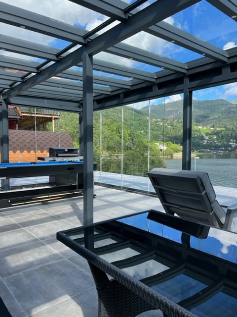 An outdoor terrace features a glass roof and partial glass walls, providing views of the surrounding landscape and water. A black glass table is accompanied by modern chairs, while a lounge chair is positioned for relaxation. A pool table is visible in the background.