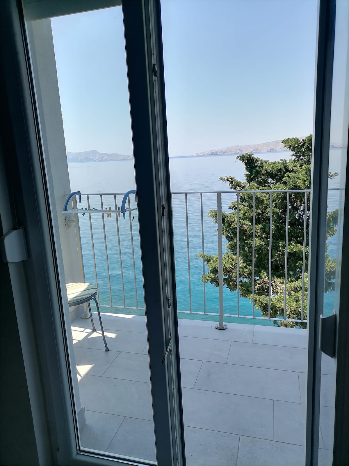 A modern balcony with a clear view of the tranquil sea, framed by sliding glass doors. A single chair is positioned next to the railing, surrounded by the natural beauty of the Mediterranean landscape.