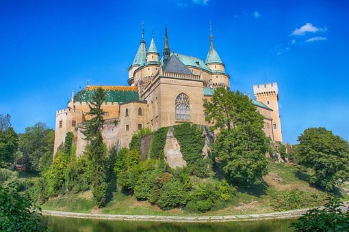 A historic stone castle is surrounded by lush greenery, featuring turrets and spires with distinctive roofs. The castle's facade displays intricate architectural details, complemented by a serene body of water that reflects its grandeur under a bright blue sky.