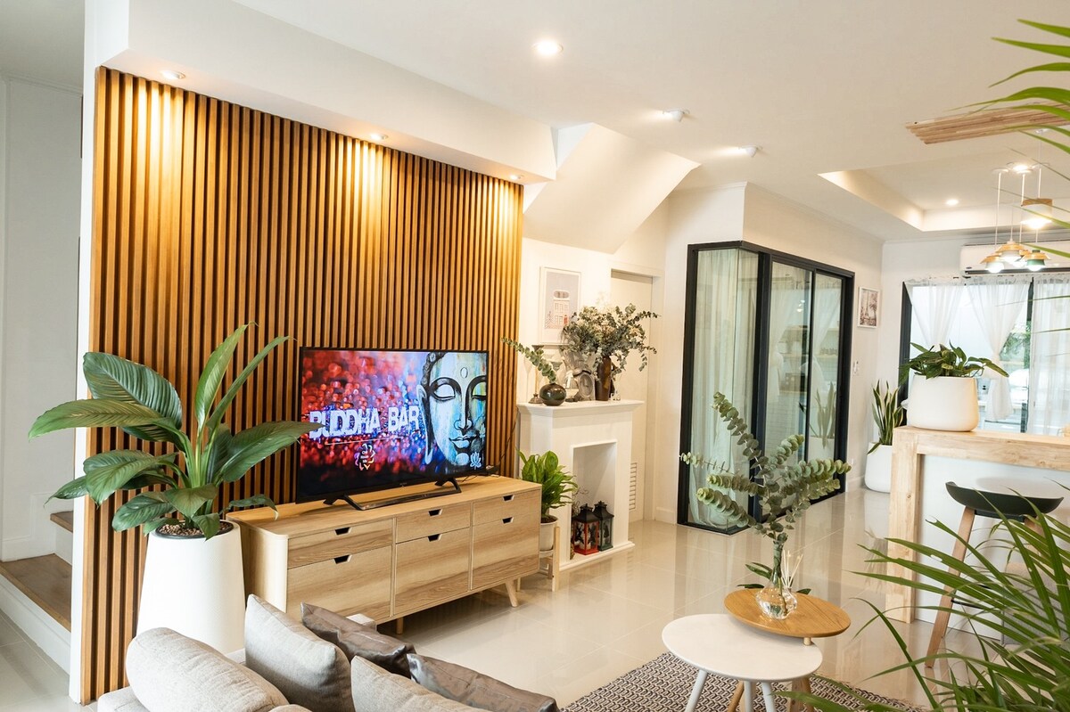 A modern living area features a wooden accent wall and a large flat-screen TV. A low wooden console holds decorative plants, while a comfortable seating arrangement includes a light-colored sofa. Large glass doors provide natural light, and indoor plants enhance the relaxed ambiance.