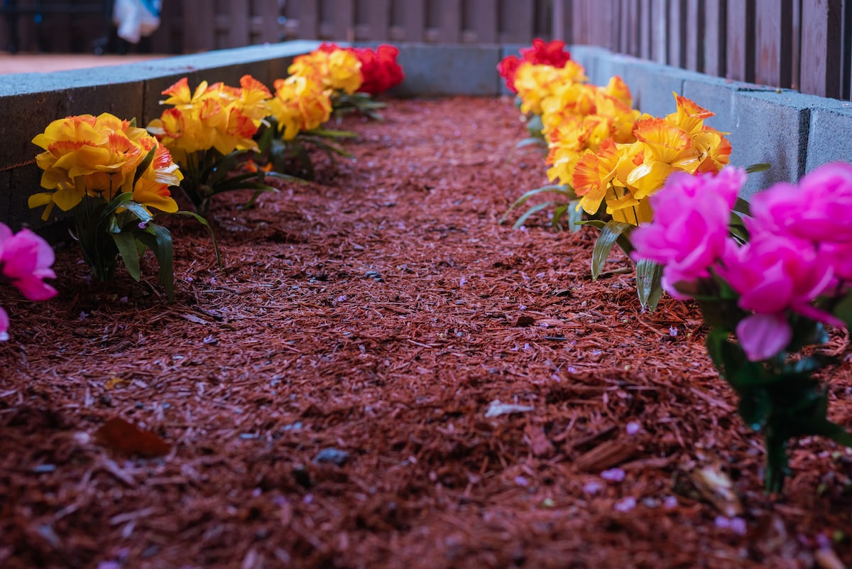 A landscaped area features vibrant artificial flowers in yellow, orange, and pink, arranged along a gravel path. The flowers are set against a backdrop of brown mulch, providing color and visual interest in the outdoor space.